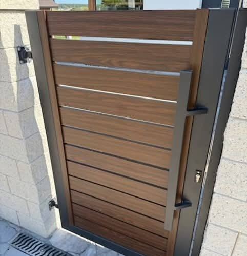 Nowoczesna furtka z poziomych desek w odcieniach brązu, z metalową ramą i długim uchwytem, zamontowana na murowanym słupku z jasnej cegły.