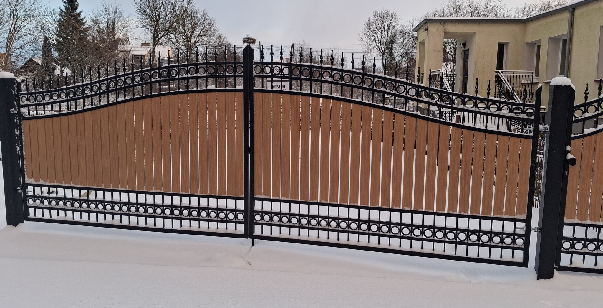 Dwuskrzydłowa brama wjazdowa z drewnianym wypełnieniem i metalową, dekoracyjną konstrukcją w śnieżnej scenerii. Widoczne budynki w tle.