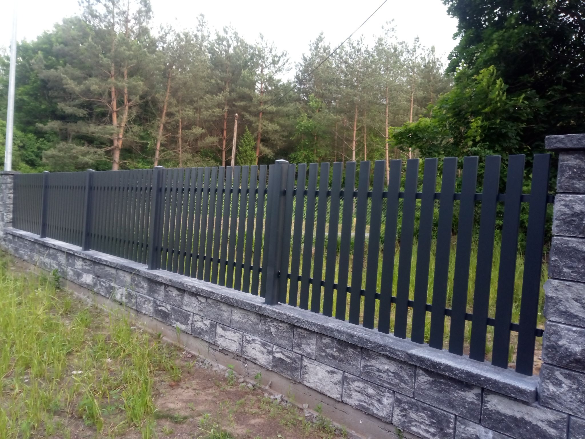 Nowoczesne, grafitowe ogrodzenie panelowe na podmurówce z szarej kostki, otoczone zielenią. Słupki ogrodzeniowe z kamienną fakturą. Widok na las w tle.