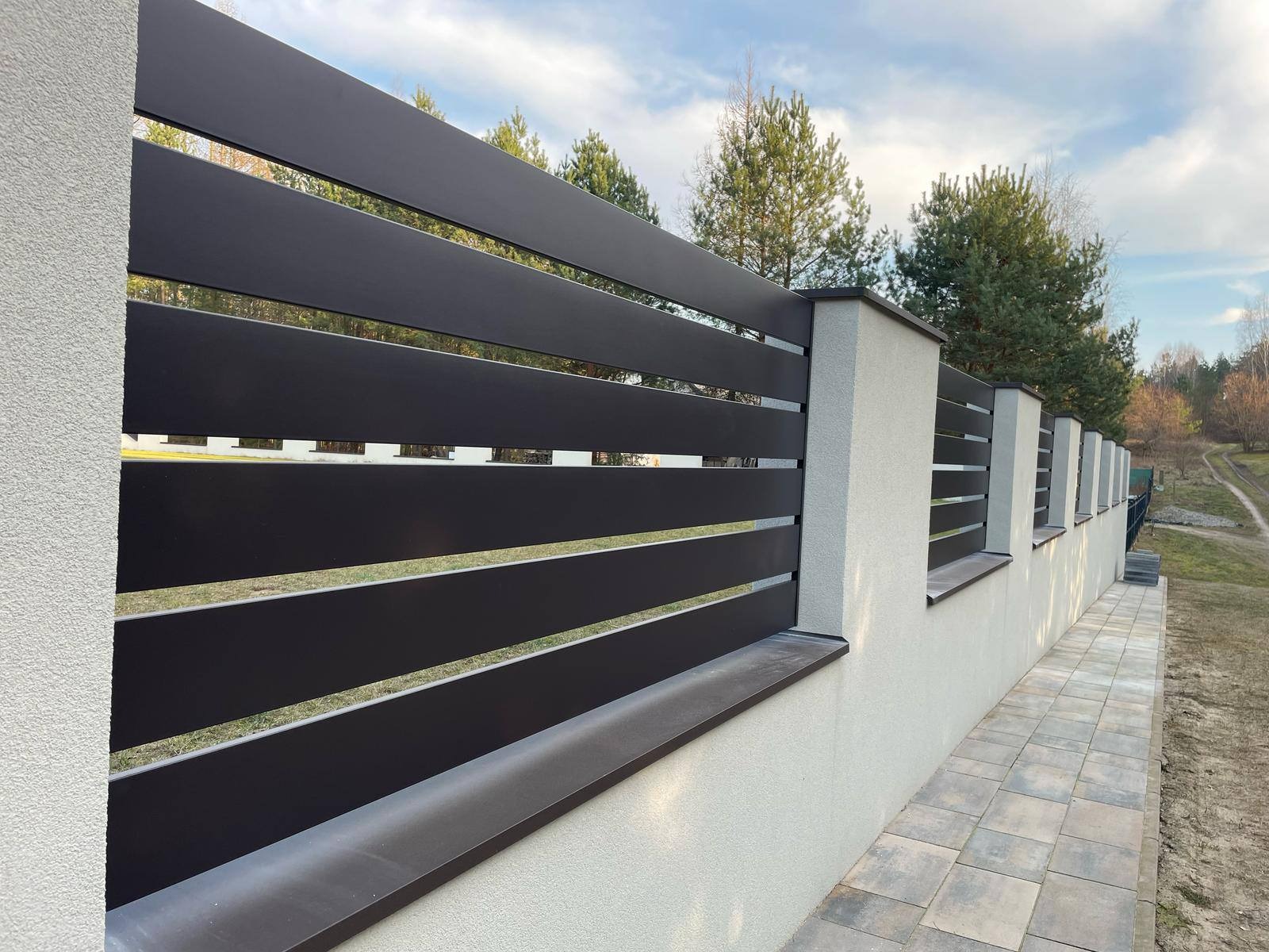 Nowoczesne ogrodzenie z poziomych, ciemnych paneli metalowych i jasnych, teksturowanych słupków. Widok z boku, z fragmentem chodnika z kostki brukowej.