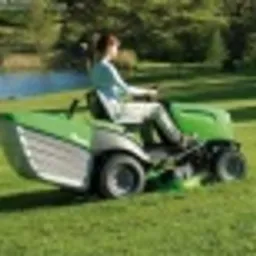 Kobieta na zielonej kosiarce traktorowej z pełnym koszem tnącą krótko przystrzyżony trawnik w słoneczny dzień. W tle drzewa i krzewy.