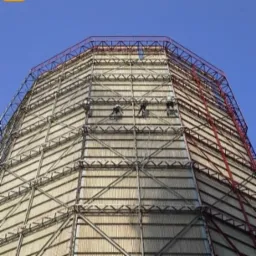 Trzech pracowników na wysokościach, zabezpieczonych linami, czyści elewację przemysłowej wieży chłodniczej o metalowej konstrukcji kratownicowej na tle błękitnego nieba.