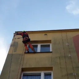 Pracownik w uprzęży alpinistycznej, zawieszony na linie, czyści elewację budynku myjką ciśnieniową. Widoczny fragment umytej ściany, okna i błękitne niebo w tle.