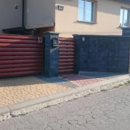 Czerwono-brązowa brama wjazdowa z poziomych desek i fragment ogrodzenia z szarych bloczków betonowych i poziomych paneli drewnianych, skrzynka na listy wbudowana w słupek.