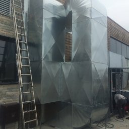 Newir Sp.zo.o - Zewnętrzny system kanałów wentylacyjnych z blachy ocynkowanej, z widoczną drabiną opartą o ścianę budynku i pracownikiem wewnątrz instalacji.