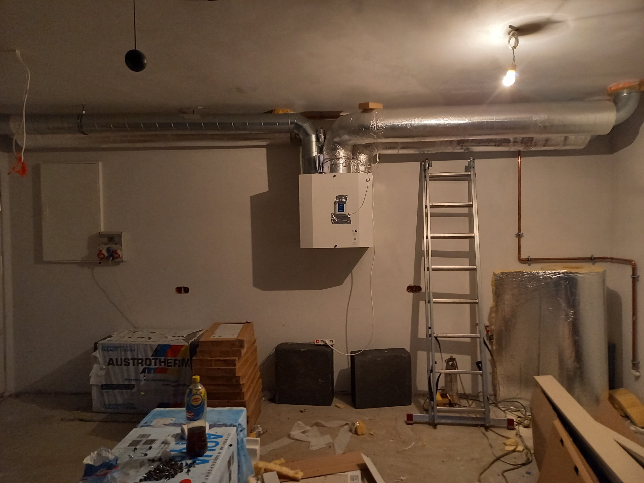 Piwnica w trakcie adaptacji: widoczne rury wentylacyjne pod sufitem, centrala wentylacyjna na ścianie, drabina, materiały budowlane i instalacje hydrauliczne.