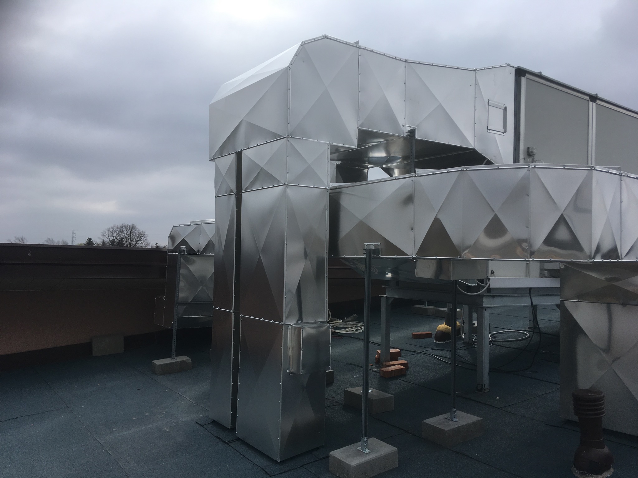 System kanałów wentylacyjnych z blachy ocynkowanej na dachu budynku, widoczne cegły i kable, pochmurne niebo w tle.