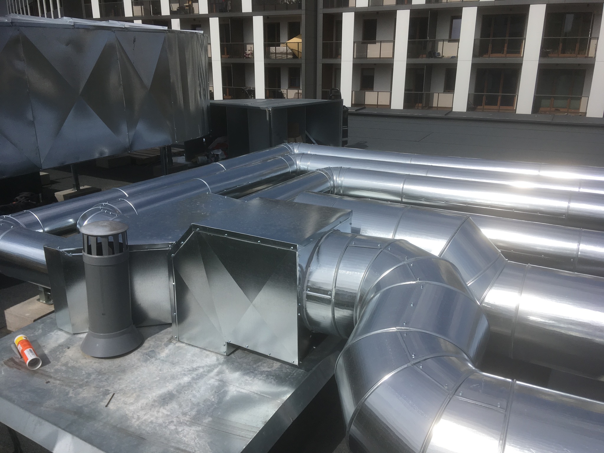 System wentylacji z błyszczących, metalowych rur na dachu budynku mieszkalnego, widoczna puszka z farbą w lewym dolnym rogu.