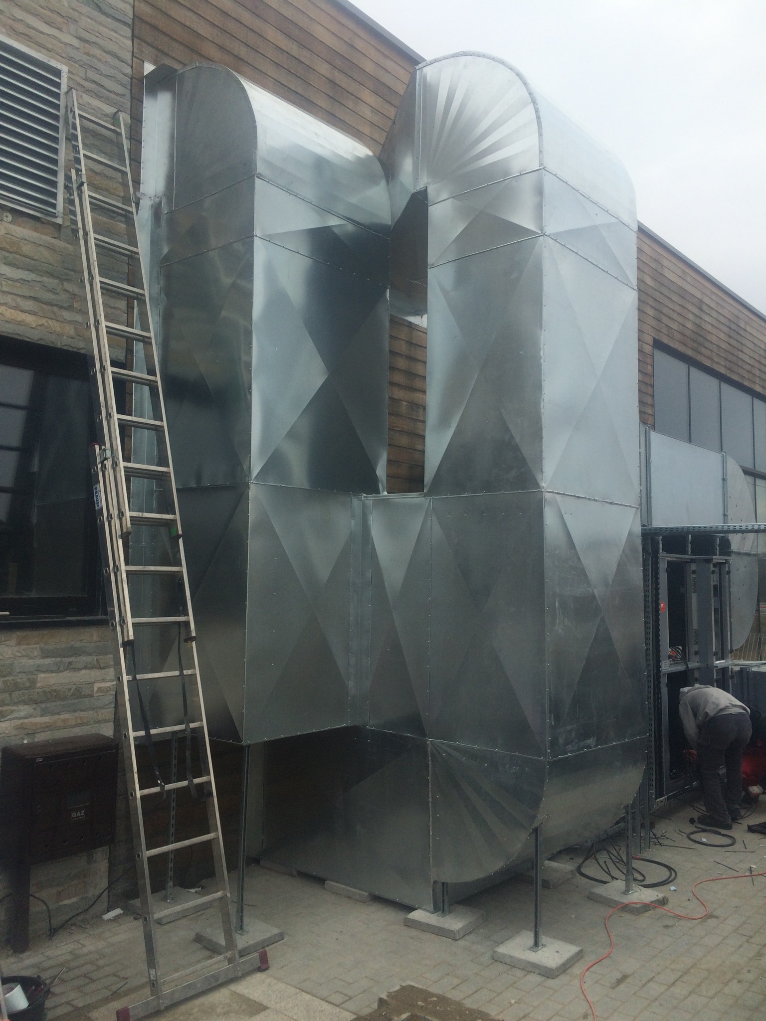 Zewnętrzny system kanałów wentylacyjnych z blachy ocynkowanej, z widoczną drabiną opartą o ścianę budynku i pracownikiem wewnątrz instalacji.