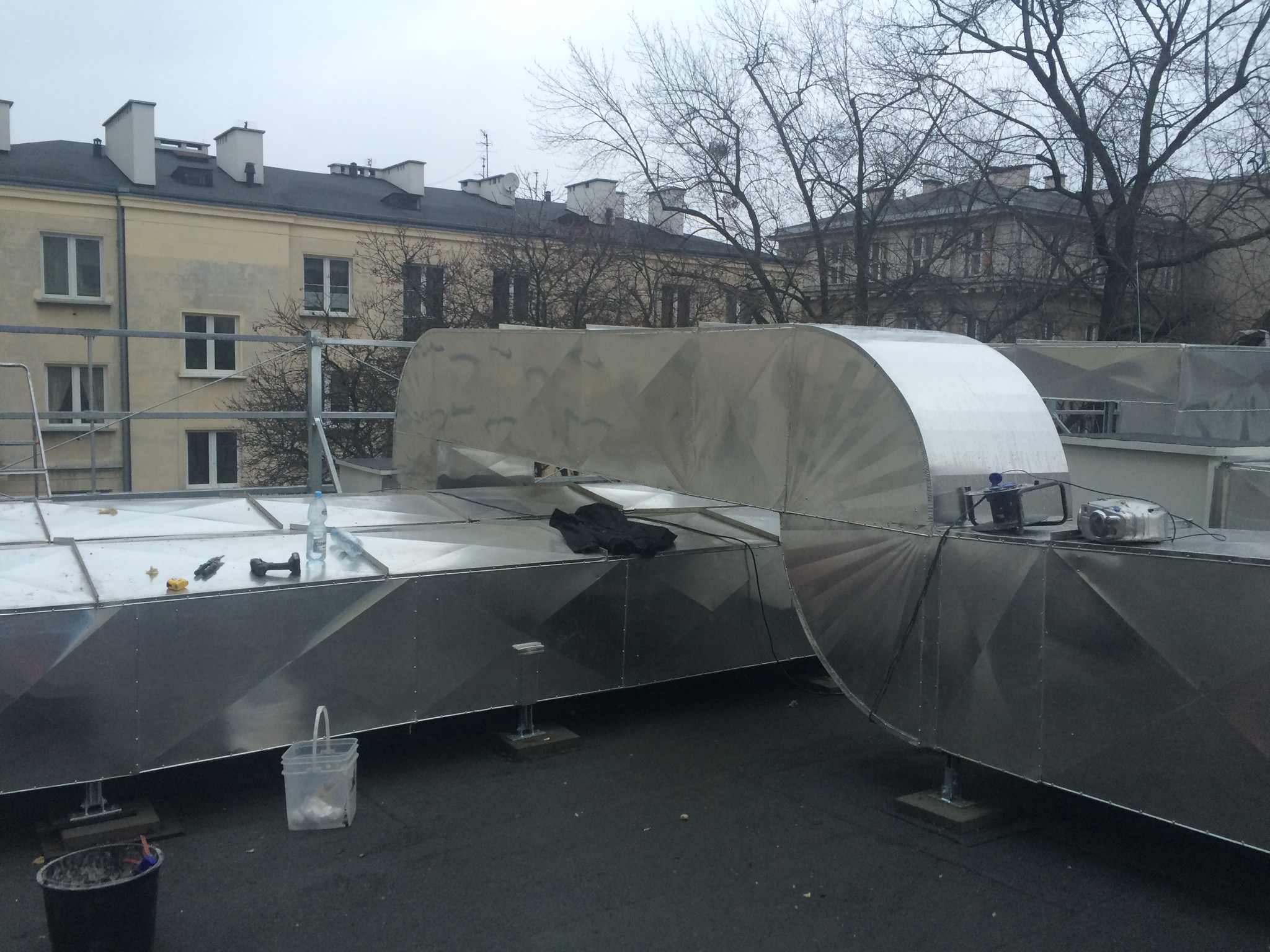 Metalowe kanały wentylacyjne na dachu budynku mieszkalnego, widoczne narzędzia, rękawice i butelka wody, pochmurne niebo w tle.