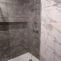 Nowoczesny prysznic z szarą, strukturalną okładziną ścienną i białą, marmurkową ścianą, oddzielony szklaną kabiną, z widocznym odpływem liniowym i chromowaną armaturą.