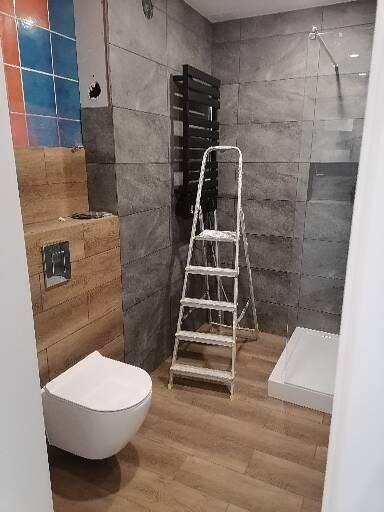 Remontowana łazienka z płytkami imitującymi kamień, wiszącą toaletą, grzejnikiem drabinkowym i prysznicem typu walk-in, widoczna drabina malarska.