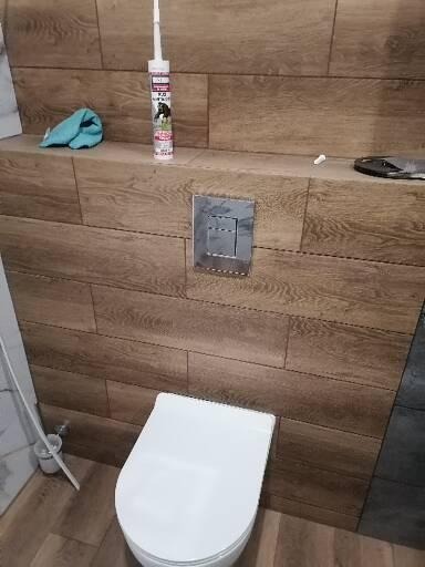 Nowoczesna toaleta z deską sedesową i przyciskiem spłukiwania na drewnopodobnej ścianie z płytek, obok leży tuba silikonu i ściereczka.