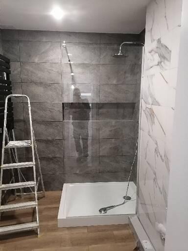 Montaż kabiny prysznicowej z szarymi płytkami imitującymi kamień na ścianie i marmurowym wykończeniem, widoczna drabina malarska i odbicie osoby w szklanej ściance.