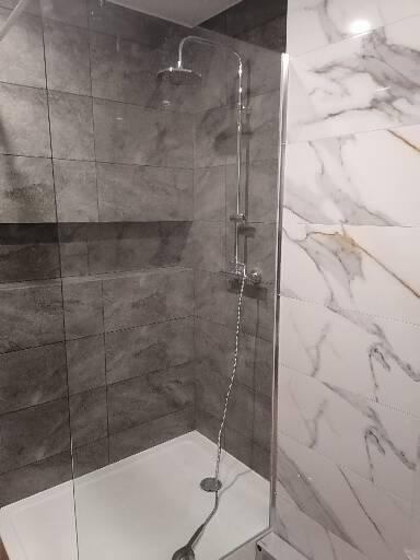 Nowoczesny prysznic z szarą, strukturalną okładziną ścienną i białą, marmurkową ścianą, oddzielony szklaną kabiną, z widocznym odpływem liniowym i chromowaną armaturą.