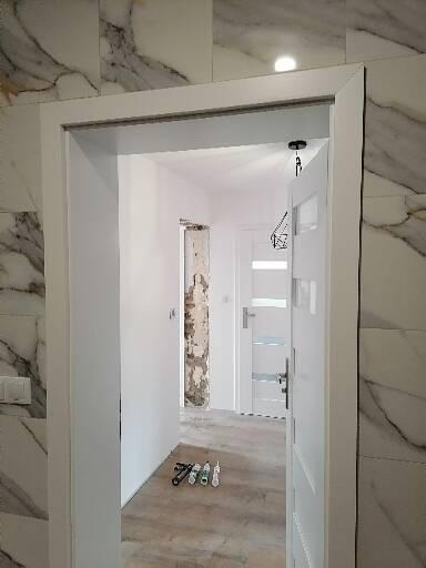 Montaż ościeżnicy w nowym otworze drzwiowym, widoczne narzędzia i białe płytki imitujące marmur.