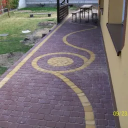 Ułożona ścieżka z kostki brukowej w kolorze bordowym z żółtymi akcentami tworzącymi wzór koła i falistej linii, biegnąca wzdłuż ściany budynku, z trawnikiem i meblami ogrodowymi w tle.