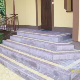 Zewnętrzne schody wejściowe wyłożone kostką brukową w odcieniach szarości i żółci, z metalową barierką po lewej stronie i brązowymi drzwiami wejściowymi w tle.