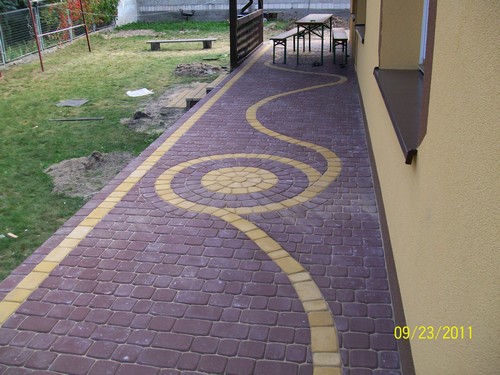 Ułożona ścieżka z kostki brukowej w kolorze bordowym z żółtymi akcentami tworzącymi wzór koła i falistej linii, biegnąca wzdłuż ściany budynku, z trawnikiem i meblami ogrodowymi w tle.