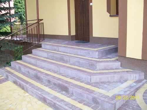 Zewnętrzne schody wejściowe wyłożone kostką brukową w odcieniach szarości i żółci, z metalową barierką po lewej stronie i brązowymi drzwiami wejściowymi w tle.