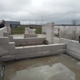 Wznoszenie ścian parteru domu z białych bloczk&oacute;w betonowych, widoczne wystające pręty zbrojeniowe oraz wiadro z zaprawą murarską na ścianie, w tle pochmurne niebo.