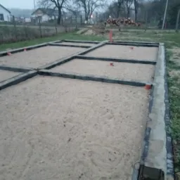 Przygotowany fundament pod dom murowany z widocznymi poziomami, zasypany piaskiem, w tle sterta drewna i budynek mieszkalny, pochmurny dzień.