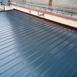 Nowoczesne, metalowe pokrycie dachowe w kolorze grafitowym, widoczne mocowania śrubowe, brązowe obrzeża dachu, tło stanowi fragment elewacji budynku z instalacją odgromową.