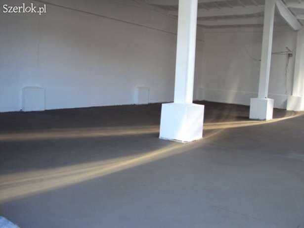 Szara, gładka powierzchnia podłogi w hali przemysłowej z białymi, kwadratowymi filarami, oświetlona przez promienie słoneczne wpadające z boku.