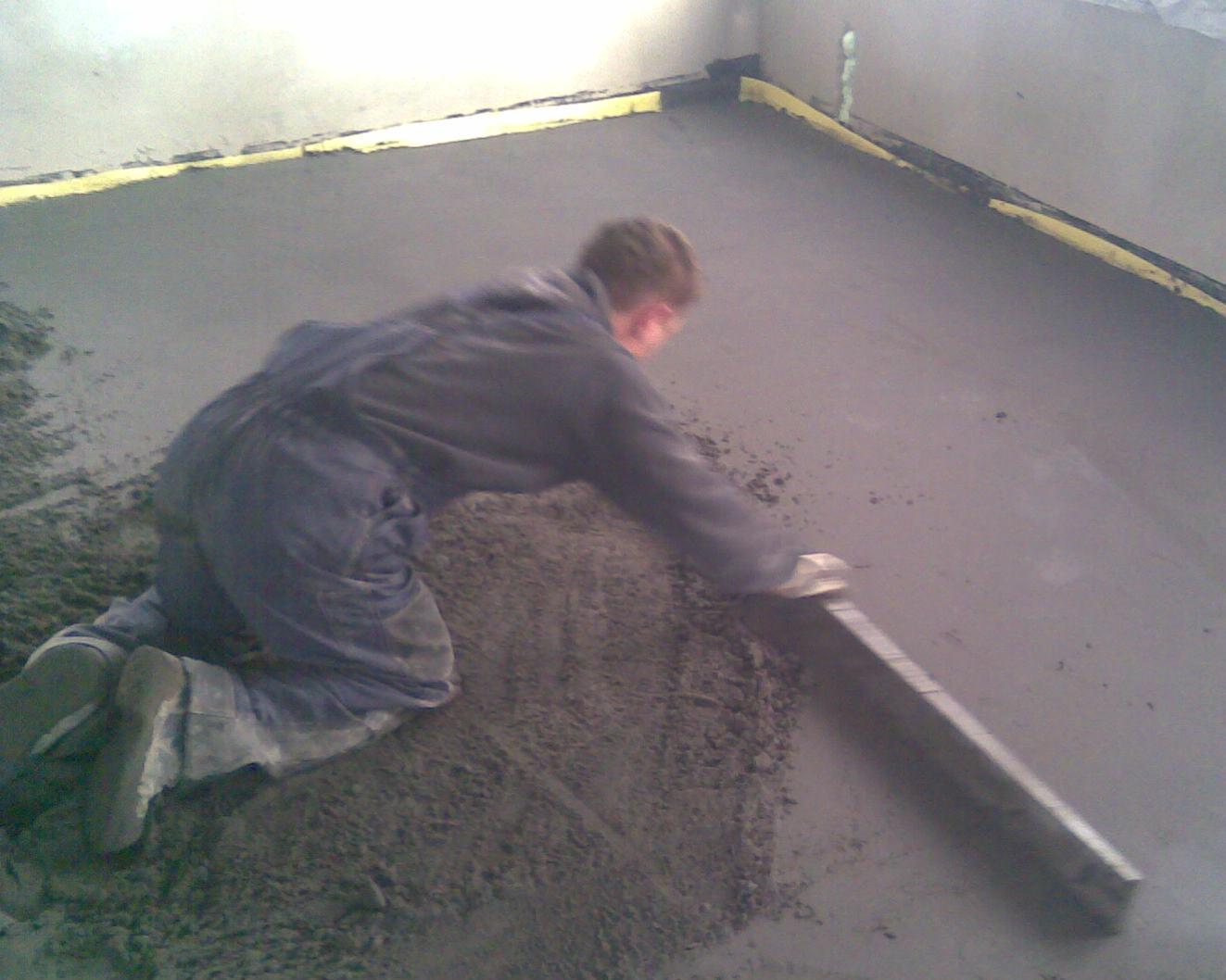 Pracownik w kombinezonie wyrównuje świeżo wylaną, szarą masę na podłodze za pomocą długiej, metalowej łaty; widoczne żółte taśmy przy ścianach.