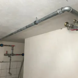 Szare rury instalacji centralnego ogrzewania i wodnej, zamontowane pod sufitem w budynku mieszkalnym z fragmentem wykończonej ściany z białymi płytkami.