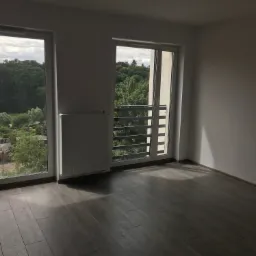 Puste, jasne wnętrze mieszkania z szarymi panelami podłogowymi, białymi ścianami, grzejnikiem i dwoma oknami z widokiem na zieleń.