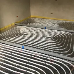 Białe rury systemu ogrzewania podłogowego ułożone spiralnie na folii z czerwoną siatką, przymocowane czarnymi klipsami, z widoczną żółtą taśmą izolacyjną przy ścianie i nieotynkowaną ścianą w tle.