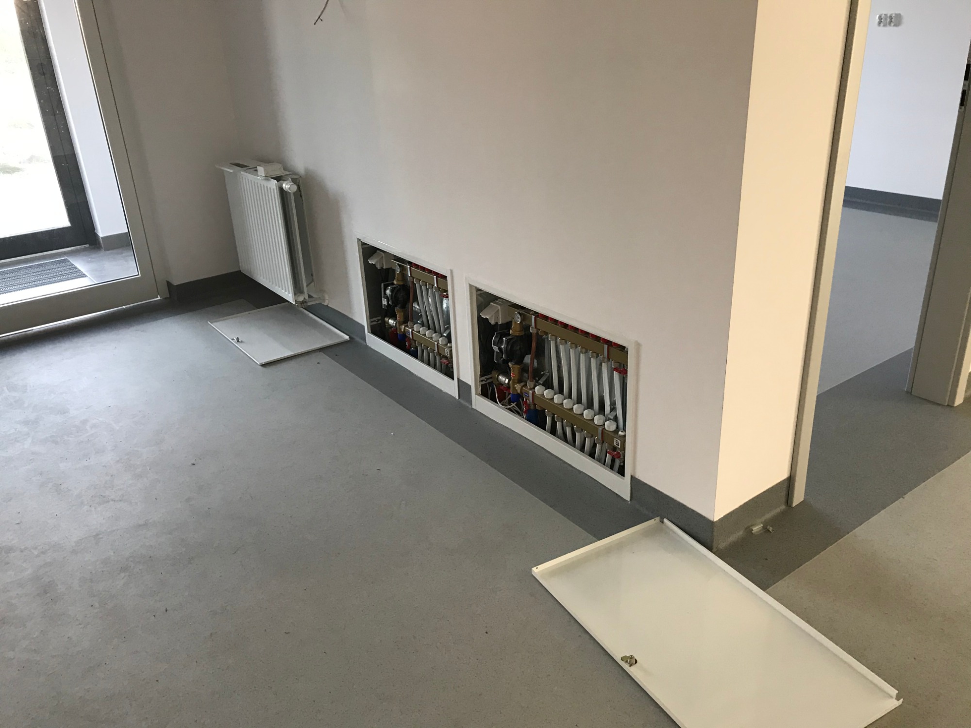 Wnętrze budynku wielorodzinnego w trakcie prac instalacyjnych: widoczne rozdzielacze ogrzewania podłogowego w dwóch wnękach, grzejnik oraz betonowa posadzka.