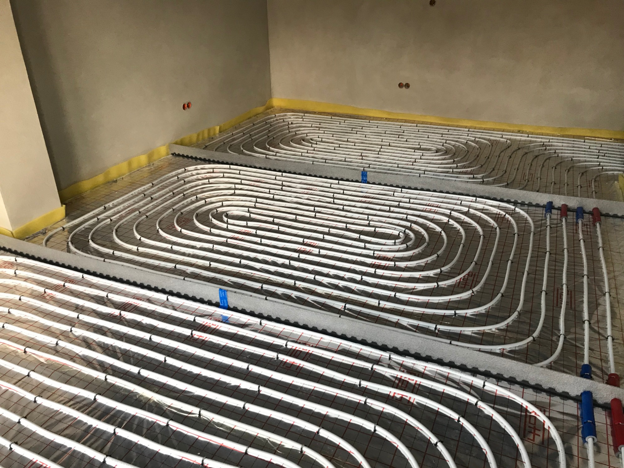 Białe rury systemu ogrzewania podłogowego ułożone spiralnie na folii z czerwoną siatką, przymocowane czarnymi klipsami, z widoczną żółtą taśmą izolacyjną przy ścianie i nieotynkowaną ścianą w tle.