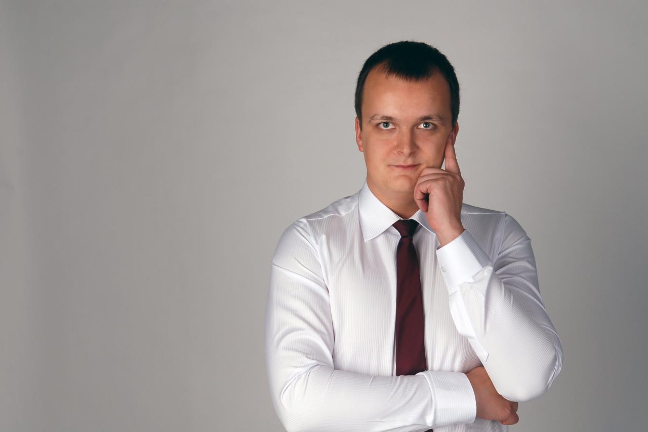 Portret mężczyzny w białej koszuli i bordowym krawacie, zamyślony, z ręką przy twarzy na jasnoszarym tle. Ujęcie od pasa w górę, z przestrzenią po lewej stronie.