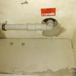 Szara rura kanalizacyjna zamontowana pod sufitem, w tle fragment ściany z płytą gipsowo-kartonową z wyciętymi otworami, widoczny fragment naklejki firmowej na suficie.