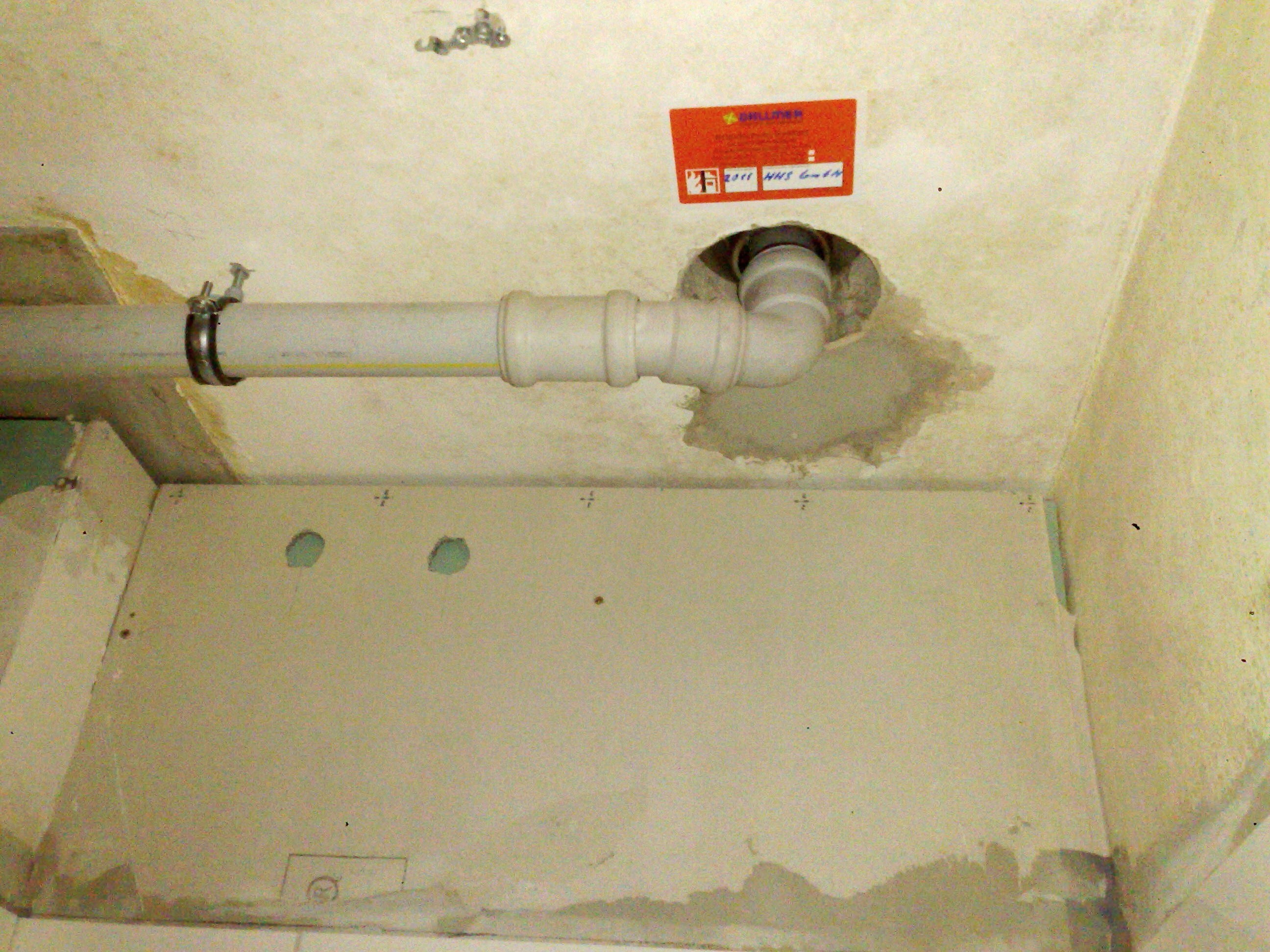 Szara rura kanalizacyjna zamontowana pod sufitem, w tle fragment ściany z płytą gipsowo-kartonową z wyciętymi otworami, widoczny fragment naklejki firmowej na suficie.