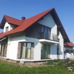 Dwukondygnacyjny dom jednorodzinny z czerwonym dachem i białymi ścianami z elementami dekoracyjnymi imitującymi drewno, balkon z balustradą ze szkła.