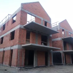 Surowy stan budynku wielorodzinnego z czerwonej cegły, widoczne betonowe balkony i niezadaszone otwory okienne, w tle inny budynek.