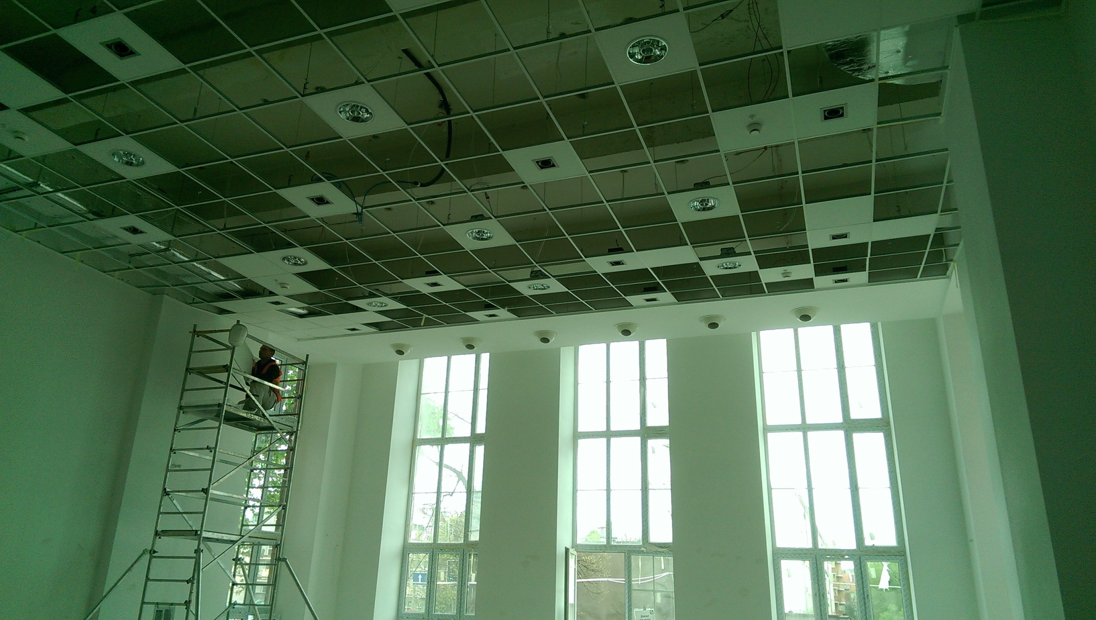 Montaż sufitu podwieszanego w biurze z widocznym stelażem, oświetleniem i pracownikiem na rusztowaniu, wysokie okna w tle.