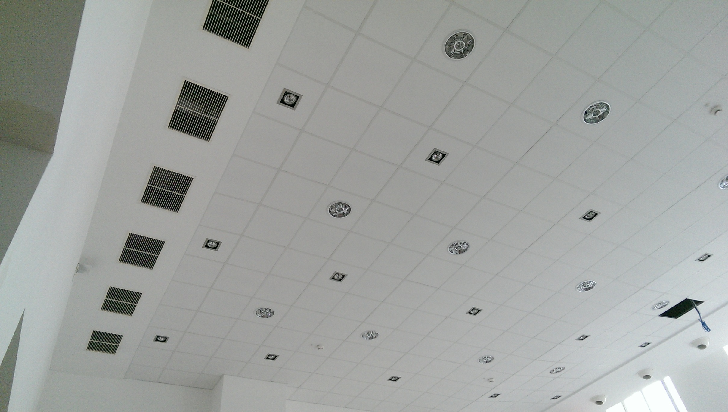 Biały sufit podwieszany z rastrowymi kratkami wentylacyjnymi i okrągłymi oprawami oświetleniowymi, widoczne także kwadratowe reflektory i czujnik dymu.