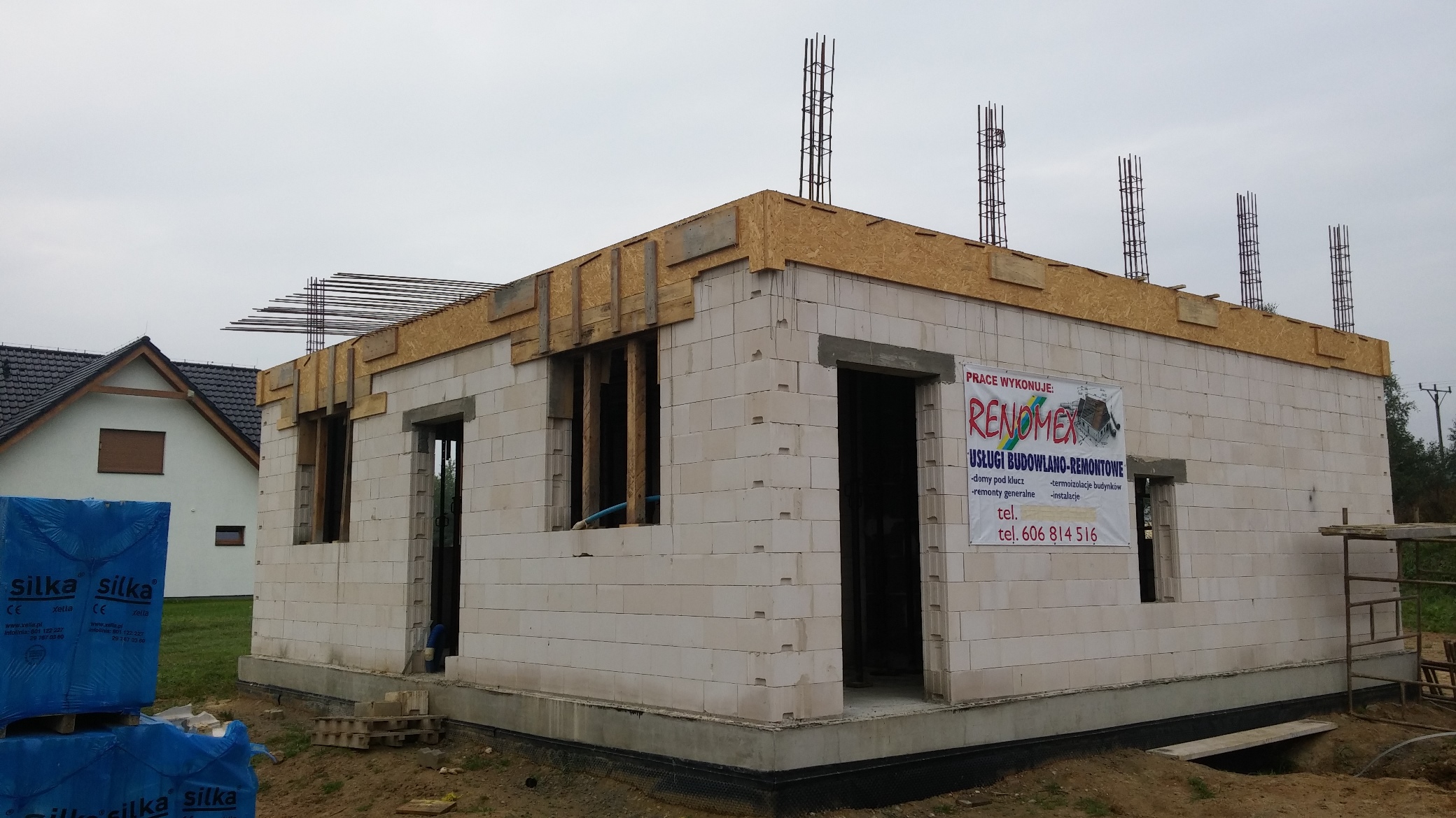 Surowy stan budowy domu jednorodzinnego z białych bloczków, widoczne zbrojenia pionowe i poziome, na ścianie baner reklamowy firmy budowlanej, w tle drugi dom.