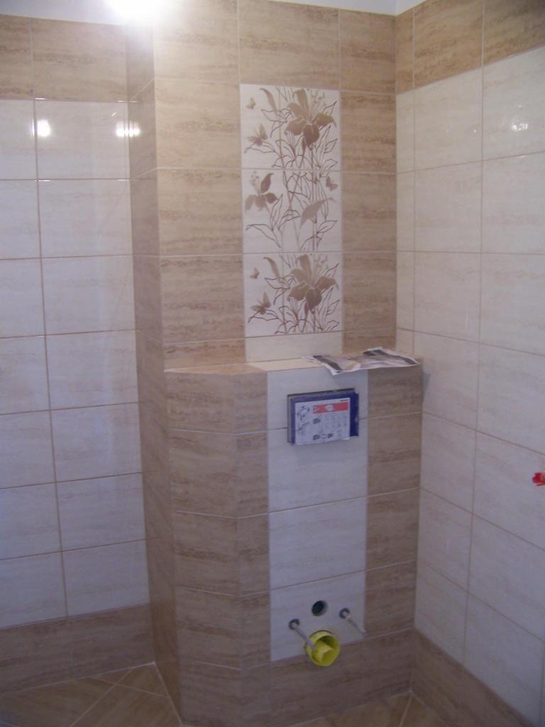 Wykończona łazienka z beżowymi płytkami imitującymi trawertyn, dekoracyjną wstawką z motywem kwiatowym i widoczną instalacją podtynkową WC.