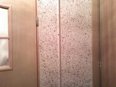 Fragment ściany pokrytej tapetą natryskową o jasnobeżowym odcieniu z drobnymi, ciemnymi plamkami, sąsiadującej z drewnianą ścianą i drzwiami z małym okienkiem.