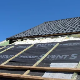 Fragment dachu w trakcie prac izolacyjnych, widoczna biała pianka między krokwiami, czarna membrana dachowa z napisem Delta Vent S oraz fragment ułożonej dachówki ceramicznej w kolorze grafitowym...