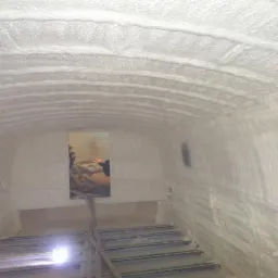 Wnętrze pomieszczenia z łukowatym sufitem i ścianami pokrytymi grubą warstwą białej pianki PUR, widoczne metalowe elementy konstrukcyjne podłogi oraz otwór w ścianie z widocznym fragmentem wnętrza...