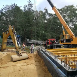 Fundamenty domu w trakcie budowy: widoczne zbrojenia, deskowanie, izolacja pionowa, koparka CAT i dźwig na tle lasu.