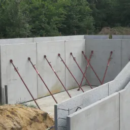 Wznoszenie betonowych ścian fundamentowych z użyciem rozpór, widoczne zbrojenia i otaczający teren budowy.