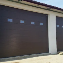 Dwie brązowe segmentowe bramy z czterema małymi kwadratowymi okienkami, zamontowane w jasnym budynku o płaskim dachu z widocznymi elementami konstrukcyjnymi.