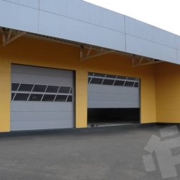 Mariusz Soszyński Przedsiębiorstwo Handlowo Usługowe - Szara brama przemysłowa z oknami częściowo otwarta w żółto-szarym budynku przemysłowym, widoczny fragment wnętrza.